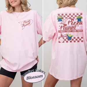 May include: Light pink t-shirt with the Pizza Planet logo on the front and back. The back features a red and white checkered design with cartoon aliens and text that reads "Pizza Planet" and "Friday Night Kids Night."