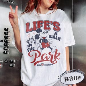 May include: White t-shirt with a graphic of Mickey Mouse walking in front of a castle, with the text "LIFE'S A WALK IN THE PARK" in red and blue. The shirt also has the text "Walt Disney World" and "Comfort Colors White".