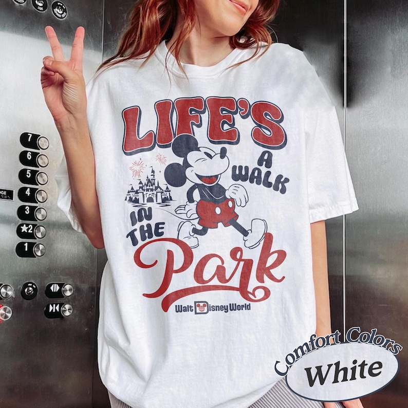 May include: White t-shirt with a graphic of Mickey Mouse walking in front of a castle, with the text "LIFE'S A WALK IN THE PARK" in red and blue. The shirt also has the text "Walt Disney World" and "Comfort Colors White".