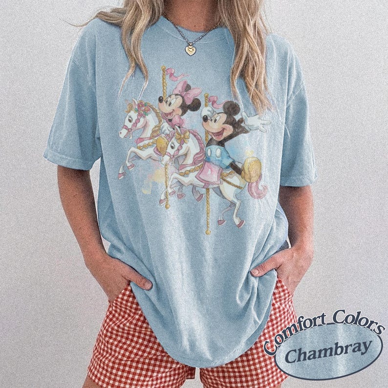 Puede incluir: Camiseta azul claro con un dise&ntilde;o de carrusel con Mickey y Minnie Mouse montados en caballos blancos. La camiseta tiene un corte holgado y mangas cortas. El texto "Comfort Colors Chambray" est&aacute; impreso en la parte inferior derecha.