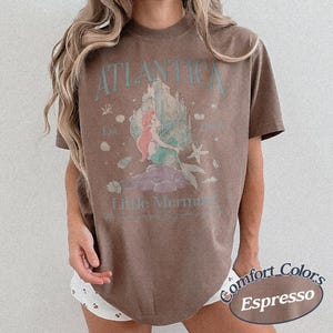 Może przedstawiać: Brązowa koszulka w kolorze espresso z grafiką syreny i napisami "Atlantica" i "Little Mermaid". Projekt zawiera zamek, muszle i rozgwiazdę. Koszulka jest częścią kolekcji Comfort Colors.