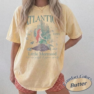 Może przedstawiać: Jasnoż&oacute;łta koszulka Comfort Colors z grafiką Małej Syrenki. Projekt zawiera słowa "ATLANTIC", "1989" i "Little Mermaid". Koszulka zawiera r&oacute;wnież frazę "We have gadgets & gizmos aplenty".