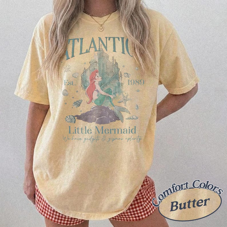Może przedstawiać: Jasnoż&oacute;łta koszulka Comfort Colors z grafiką Małej Syrenki. Projekt zawiera słowa "ATLANTIC", "1989" i "Little Mermaid". Koszulka zawiera r&oacute;wnież frazę "We have gadgets & gizmos aplenty".