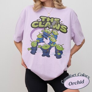 May include: A light purple t-shirt with a graphic of green aliens in blue uniforms and the text "THE CLAWS". The design includes a purple ring and a claw machine. The shirt is labeled "Comfort Colors Orchid".