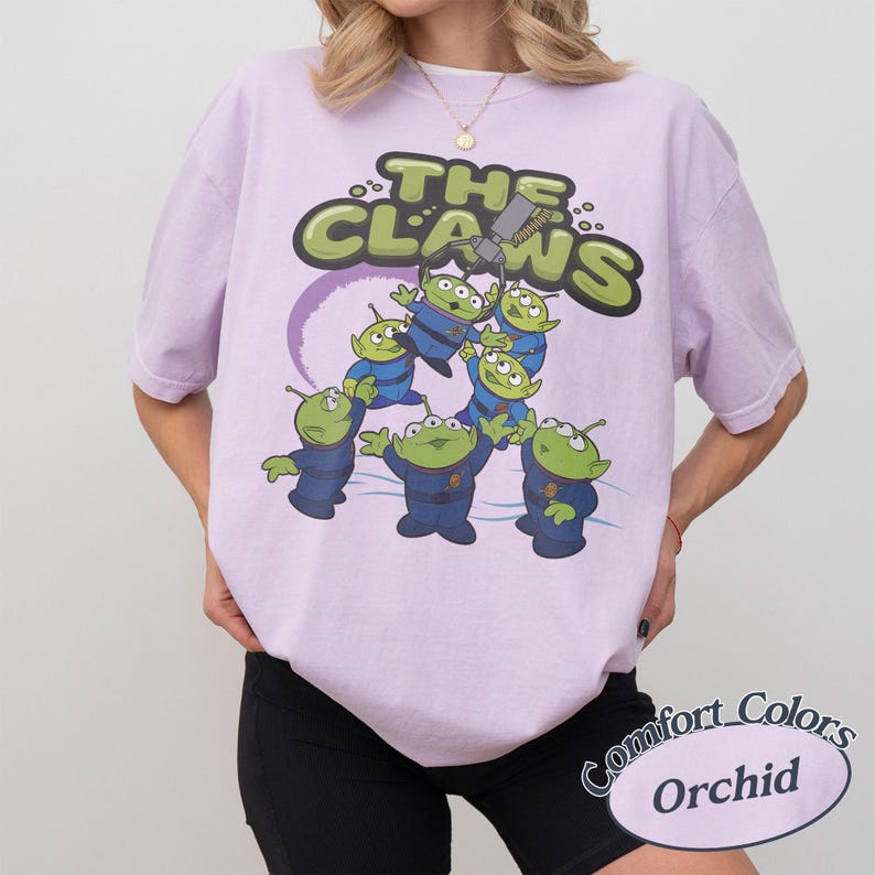 May include: A light purple t-shirt with a graphic of green aliens in blue uniforms and the text "THE CLAWS". The design includes a purple ring and a claw machine. The shirt is labeled "Comfort Colors Orchid".
