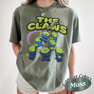 May include: Olive green t-shirt featuring the phrase "The Claws" in a playful font, with a graphic of green aliens in blue uniforms being lifted by a claw machine. The shirt is labeled "Comfort Colors Moss".