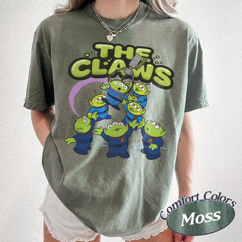 May include: Olive green t-shirt featuring the phrase "The Claws" in a playful font, with a graphic of green aliens in blue uniforms being lifted by a claw machine. The shirt is labeled "Comfort Colors Moss".