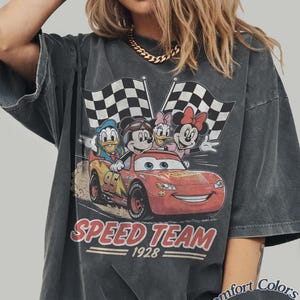 K&ouml;nnte beinhalten: Dunkelgraues T-Shirt mit einer Grafik von Mickey Mouse, Donald Duck, Daisy Duck und Minnie Mouse in einem Rennwagen mit karierten Flaggen. Die Worte "SPEED TEAM 1928" sind darunter gedruckt. Das Shirt ist mit "Comfort Colors Pepper" gekennzeichnet.