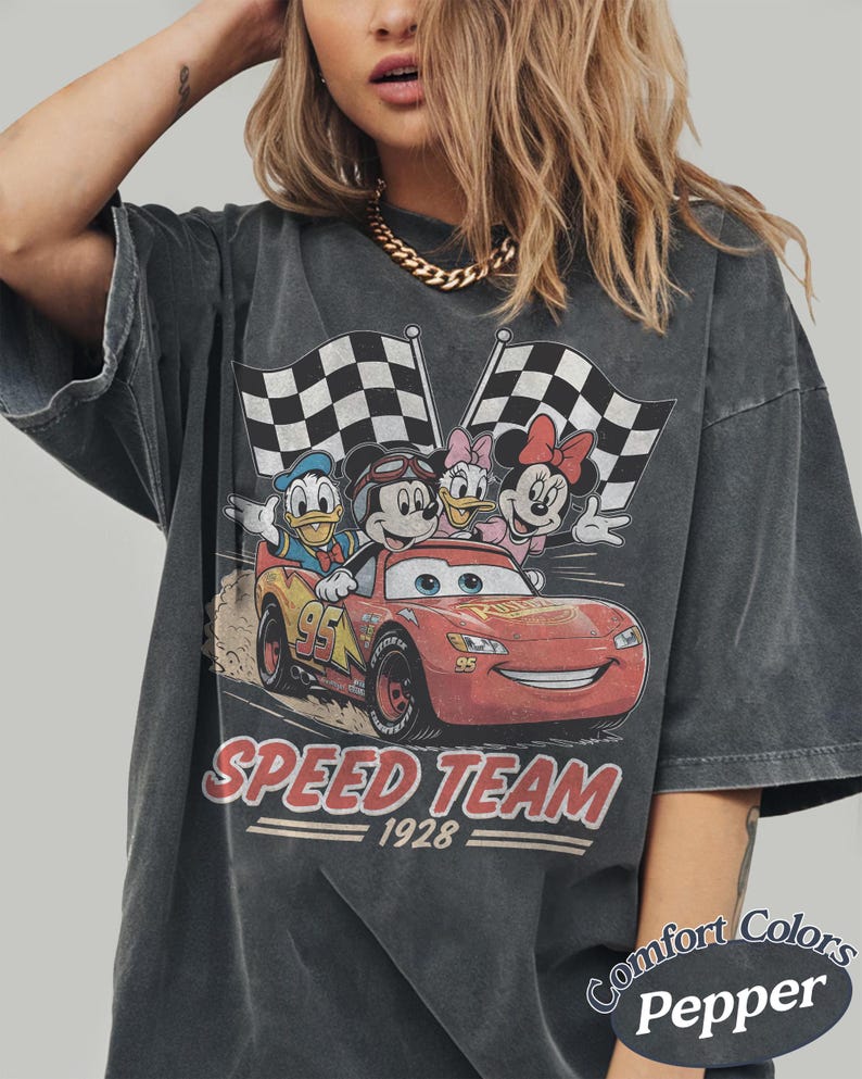 K&ouml;nnte beinhalten: Dunkelgraues T-Shirt mit einer Grafik von Mickey Mouse, Donald Duck, Daisy Duck und Minnie Mouse in einem Rennwagen mit karierten Flaggen. Die Worte "SPEED TEAM 1928" sind darunter gedruckt. Das Shirt ist mit "Comfort Colors Pepper" gekennzeichnet.