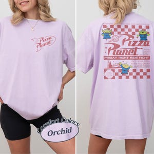 May include: Orchid-colored t-shirt with a Pizza Planet logo on the front and a graphic on the back. The back graphic features the Pizza Planet logo, aliens, and text. The shirt is a Comfort Colors brand.