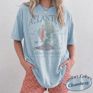Może przedstawiać: Jasnoniebieska koszulka z grafiką syreny i napisami "Atlantis" i "Little Mermaid". Koszulka ma r&oacute;wnież napis "Est. 1989". Wykonana z materiału Comfort Colors Chambray.