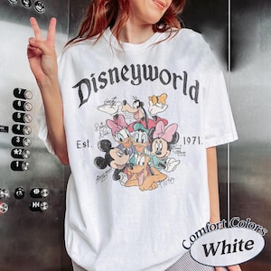 Może przedstawiać: Biała koszulka z grafiką Disneyworld z Myszka Miki, Myszka Minnie, Kaczorem Donaldem, Daisy Duck, Goofym i Pluto. Koszulka zawiera tekst "Disneyworld Est. 1971." i "Comfort Colors White."