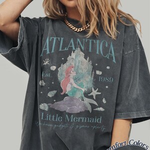 Może przedstawiać: Ciemnoszara koszulka z napisem "ATLANTICA" i "Little Mermaid", z grafiką syreny i zamku. Koszulka ma r&oacute;wnież napis "Est. 1989" i "We have gadgets & gizmos aplenty". Koszulka to Comfort Colors Pepper.