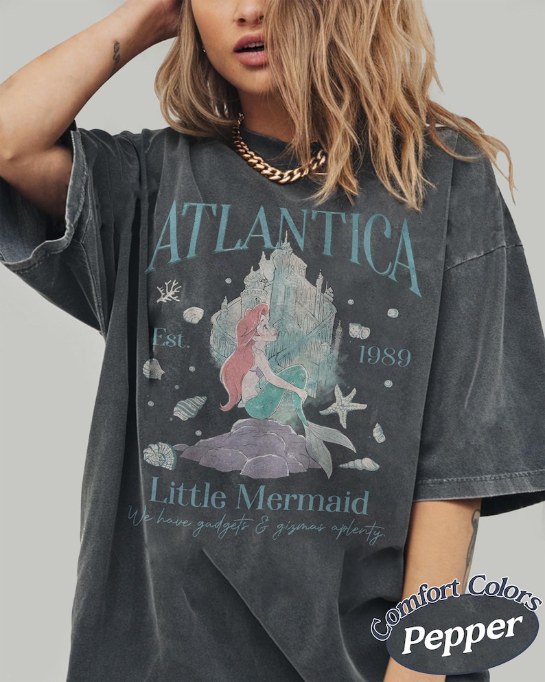 Może przedstawiać: Ciemnoszara koszulka z napisem "ATLANTICA" i "Little Mermaid", z grafiką syreny i zamku. Koszulka ma r&oacute;wnież napis "Est. 1989" i "We have gadgets & gizmos aplenty". Koszulka to Comfort Colors Pepper.