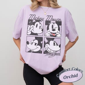 Op de afbeelding: Een lichtpaars T-shirt met een vierpaneelsafbeelding van Mickey Mouse met verschillende uitdrukkingen. Het shirt heeft de woorden "Mickey Mouse" boven de afbeelding en "Comfort Colors Orchid" eronder.