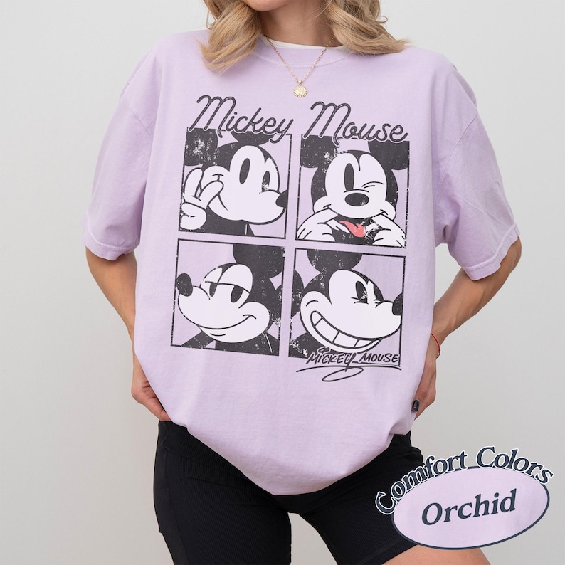Op de afbeelding: Een lichtpaars T-shirt met een vierpaneelsafbeelding van Mickey Mouse met verschillende uitdrukkingen. Het shirt heeft de woorden "Mickey Mouse" boven de afbeelding en "Comfort Colors Orchid" eronder.