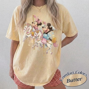 Puede incluir: Camiseta amarilla clara Comfort Colors con un dise&ntilde;o de carrusel con los personajes de Mickey y Minnie Mouse. El gr&aacute;fico muestra a los personajes montados en caballos blancos con detalles rosas y dorados. La camiseta tiene un corte holgado y el texto "Comfort Colors Butter" est&aacute; impreso abajo a la derecha.