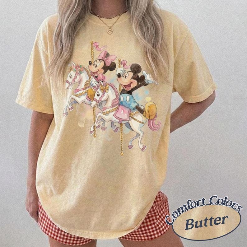 Puede incluir: Camiseta amarilla clara Comfort Colors con un dise&ntilde;o de carrusel con los personajes de Mickey y Minnie Mouse. El gr&aacute;fico muestra a los personajes montados en caballos blancos con detalles rosas y dorados. La camiseta tiene un corte holgado y el texto "Comfort Colors Butter" est&aacute; impreso abajo a la derecha.
