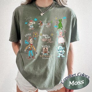 Puede incluir: Camiseta verde musgo con personajes de dibujos animados de Toy Story. El dise&ntilde;o incluye a Woody, Jessie, Slinky Dog, Rex, Bo Peep y Buzz Lightyear. El texto dice "Andy's Room Keep Out" y "Wanted". La camiseta est&aacute; etiquetada como "Comfort Colors Moss".