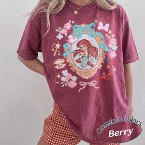 Puede incluir: Camiseta color bayas con una ilustraci&oacute;n enmarcada de una princesa pelirroja. El marco est&aacute; rodeado de flores rosas, lazos azules y otros elementos decorativos. El texto "Comfort Colors Berry" est&aacute; impreso en la parte inferior derecha.