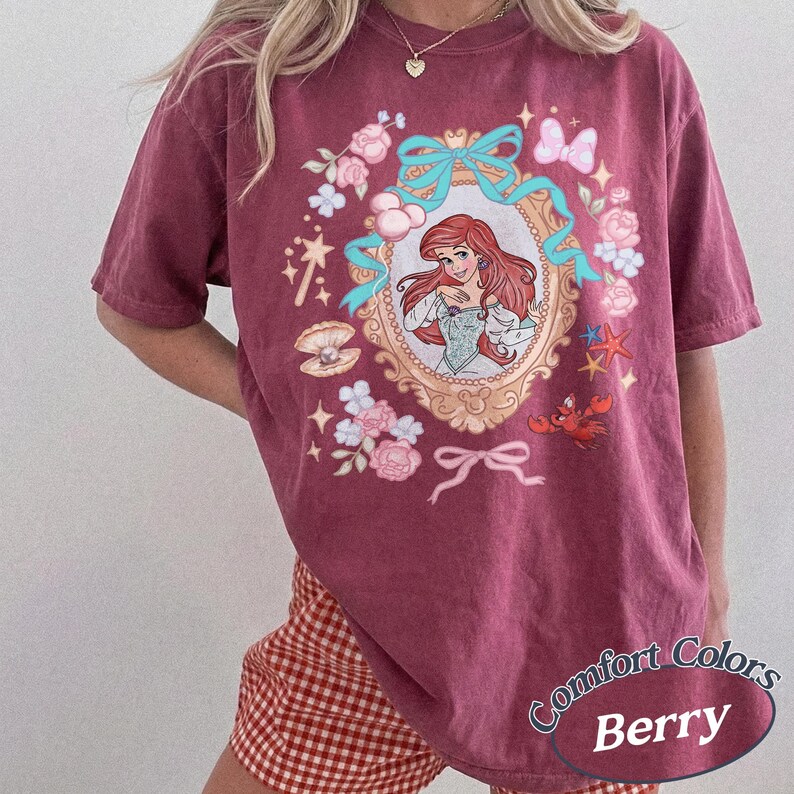 Puede incluir: Camiseta color bayas con una ilustraci&oacute;n enmarcada de una princesa pelirroja. El marco est&aacute; rodeado de flores rosas, lazos azules y otros elementos decorativos. El texto "Comfort Colors Berry" est&aacute; impreso en la parte inferior derecha.