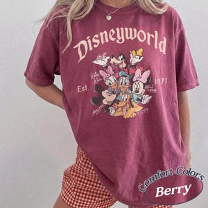 Może przedstawiać: Koszulka w kolorze jagodowym z napisem "Disneyworld" i grafiką postaci Disneya. Koszulka przedstawia Myszka Miki, Minnie, Kaczora Donalda, Daisy, Goofy'ego i Pluto. Widoczny jest r&oacute;wnież napis "Est. 1971".