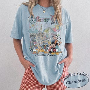 May include: Pale blue t-shirt with a Disney trip design, including Mickey Mouse and friends, fireworks, and the text "Disney Trip 2026". The shirt also features "Custom Name" and "Comfort Colors Chambray".