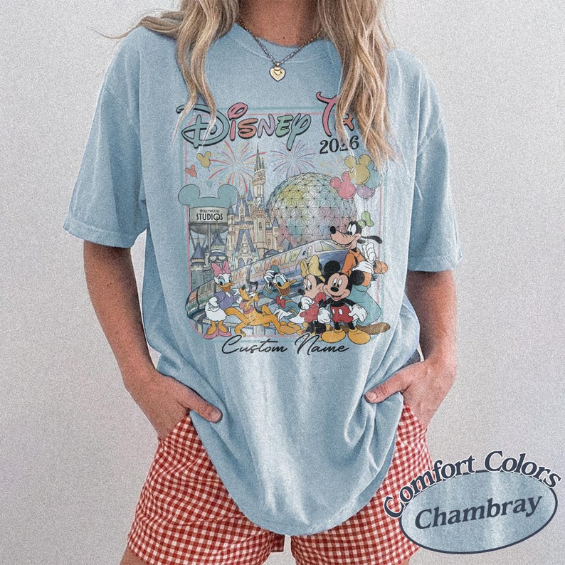 May include: Pale blue t-shirt with a Disney trip design, including Mickey Mouse and friends, fireworks, and the text "Disney Trip 2026". The shirt also features "Custom Name" and "Comfort Colors Chambray".