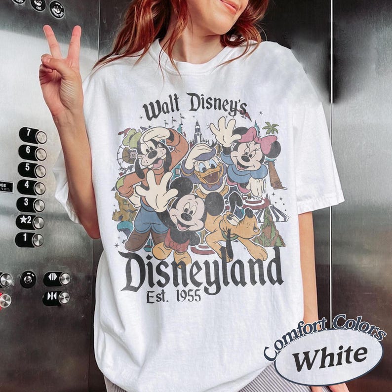 May include: White t-shirt featuring a graphic of Disney characters including Mickey Mouse, Donald Duck, and Goofy. The shirt has the text "Walt Disney's Disneyland Est. 1955" and "Comfort Colors White".