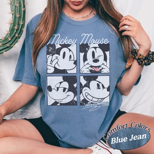Op de afbeelding: Blauw T-shirt met korte mouwen met een vierpaneelsafbeelding van Mickey Mouse met verschillende uitdrukkingen. De tekst "Mickey Mouse" staat boven de afbeelding. Het shirt is gelabeld "Comfort Colors Blue Jean".