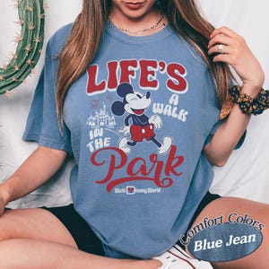 May include: Blue jean-colored t-shirt featuring a graphic of Mickey Mouse walking in front of Cinderella's castle. The text reads "Life's a Walk in the Park" and "Walt Disney World" in red and white lettering. The shirt is a Comfort Colors brand.