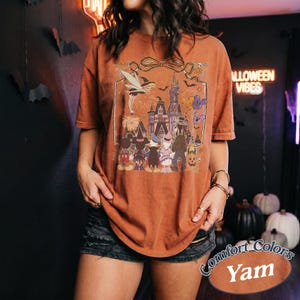 Pode incluir: Uma camiseta laranja queimado com um gr&aacute;fico de Halloween com um castelo, fogos de artif&iacute;cio e personagens de desenhos animados. A camisa tem um ajuste folgado e as palavras "Comfort Colors Yam" s&atilde;o impressas em um r&oacute;tulo.