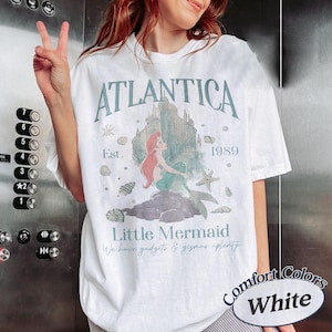 Może przedstawiać: Biała koszulka z grafiką z napisem "ATLANTICA" i "Little Mermaid". Projekt zawiera syrenę, zamek i muszle. Koszulka ma r&oacute;wnież napis "Est. 1989" i "We have gadgets & gizmas aplenty".