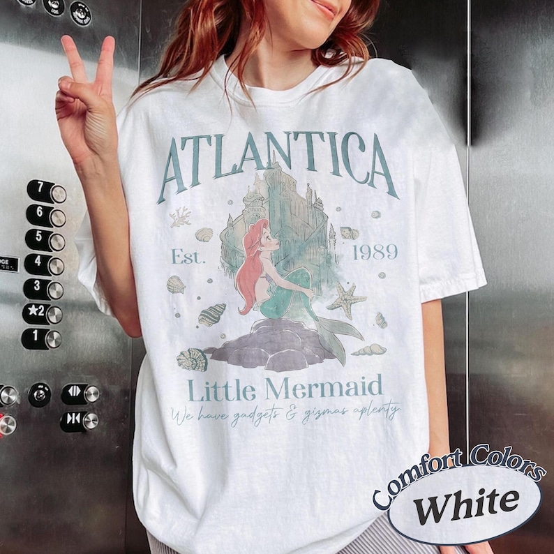 Może przedstawiać: Biała koszulka z grafiką z napisem "ATLANTICA" i "Little Mermaid". Projekt zawiera syrenę, zamek i muszle. Koszulka ma r&oacute;wnież napis "Est. 1989" i "We have gadgets & gizmas aplenty".