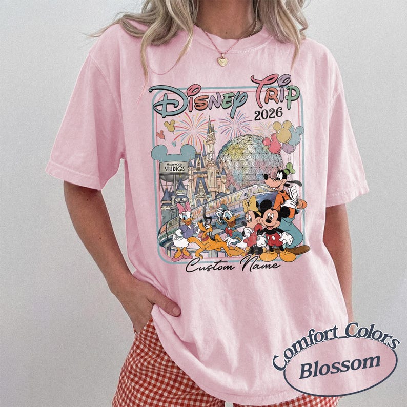 May include: A pale pink t-shirt with a vibrant graphic of Disney characters and attractions. The design includes the text "Disney Trip 2026" and "Custom Name". The shirt is labelled "Comfort Colors Blossom".