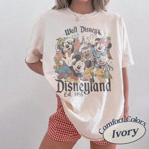 May include: Ivory-colored t-shirt featuring a vintage Disneyland graphic with Mickey Mouse, Goofy, Donald Duck, and Minnie Mouse. The shirt has the text "Walt Disney's Disneyland Est. 1955". The shirt is a Comfort Colors brand.
