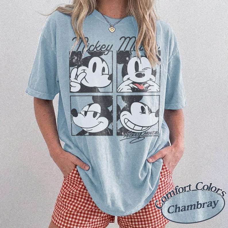 Op de afbeelding: Lichtblauw T-shirt met een vierdelige Mickey Mouse-afbeelding en de woorden "Mickey Mouse". Het shirt heeft ook de woorden "Comfort Colors Chambray". De persoon draagt een rood-witte geruite short. Het T-shirt is gemaakt van zacht materiaal.