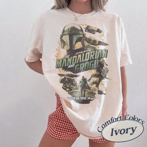 Może przedstawiać: Kremowa koszulka z grafiką z "The Mandalorian and Grogu". Projekt zawiera hełm Mandalorianina, Grogu i napis "THIS IS THE WAY". Koszulka marki Comfort Colors.