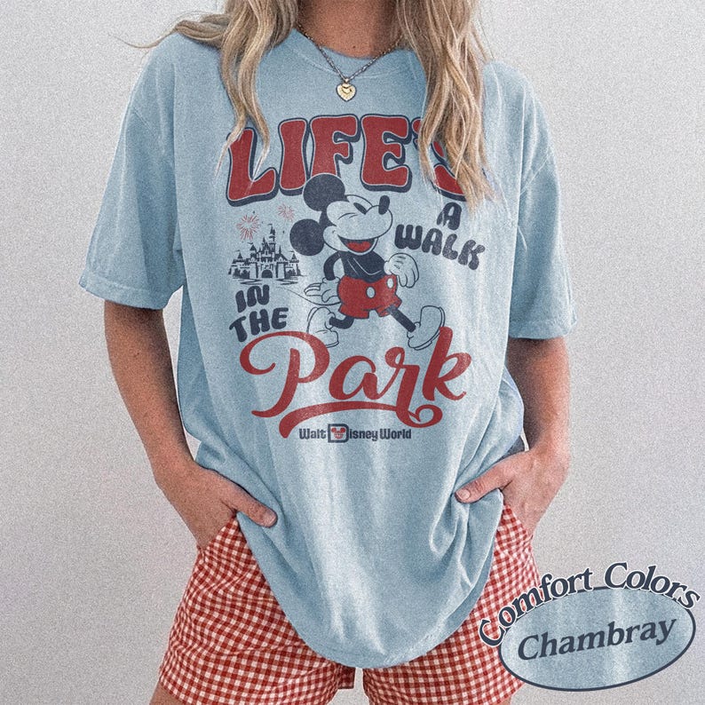 May include: Light blue t-shirt with a graphic of Mickey Mouse and the text "LIFE'S A WALK IN THE PARK" in red and white. The shirt also features the words "Walt Disney World". The shirt is a Comfort Colors Chambray.
