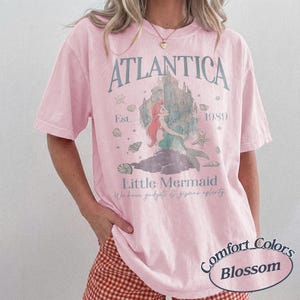 Może przedstawiać: Jasnor&oacute;żowa koszulka z grafiką syreny, napisami "ATLANTICA" i "Little Mermaid." Projekt zawiera zamek, muszle i rok "1989." Koszulka jest od Comfort Colors, Blossom.