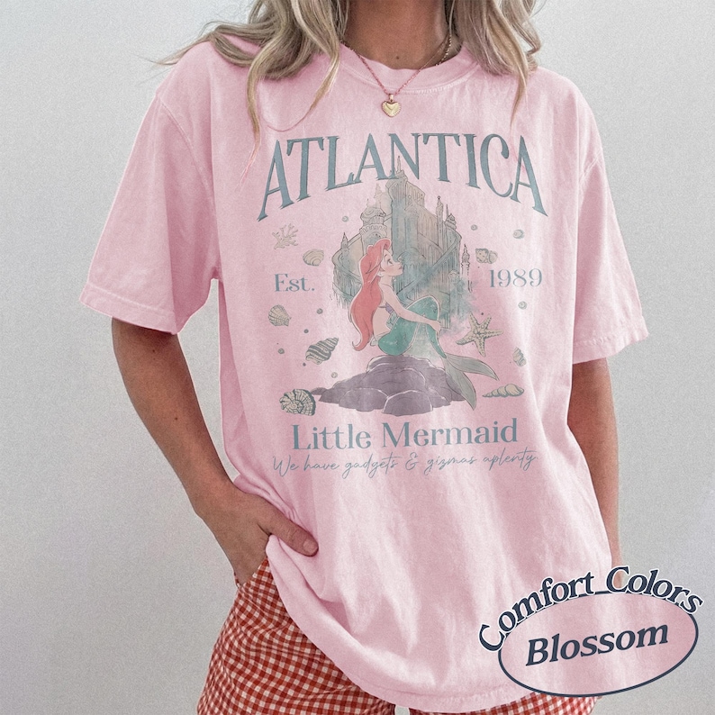 Może przedstawiać: Jasnor&oacute;żowa koszulka z grafiką syreny, napisami "ATLANTICA" i "Little Mermaid." Projekt zawiera zamek, muszle i rok "1989." Koszulka jest od Comfort Colors, Blossom.