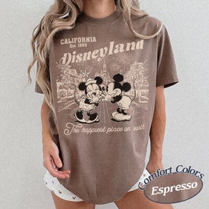 Puede incluir: Camiseta color espresso con un dise&ntilde;o vintage de Disneyland con Mickey y Minnie Mouse. El dise&ntilde;o incluye el texto "CALIFORNIA Est. 1955" y "The happiest place on earth."