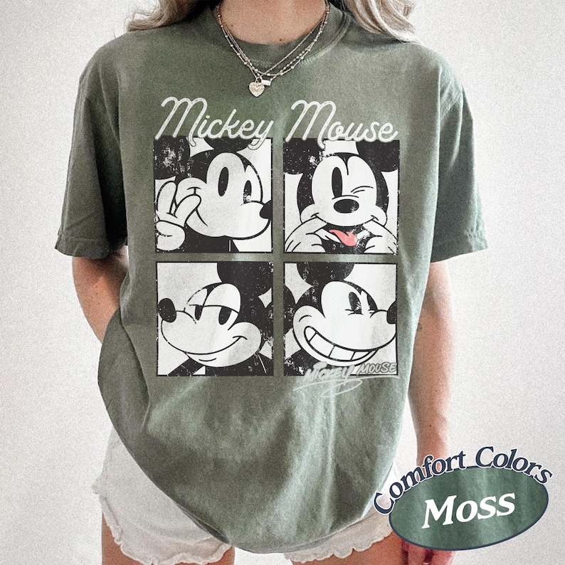 Op de afbeelding: Olijfgroen t-shirt met een vier-paneel ontwerp van Mickey Mouse met verschillende uitdrukkingen. De woorden "Mickey Mouse" staan in een scriptletter boven de panelen. Het shirt is gelabeld "Comfort Colors Moss".