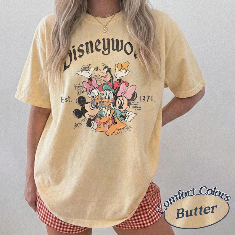 Może przedstawiać: Jasnoż&oacute;łta koszulka z grafiką postaci Disneya, w tym Myszki Miki i Myszki Minnie, Kaczora Donalda i Goofy'ego. Koszulka ma napis "Disneyworld Est. 1971" w stylu vintage. Koszulka to Comfort Colors Butter.