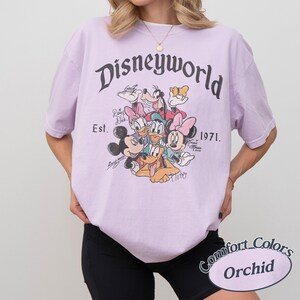 Może przedstawiać: Jasnofioletowa koszulka z napisem "Disneyworld" w ozdobnej czcionce, z grafiką postaci Disneya. Koszulka zawiera napis "Est. 1971." i logo Comfort Colors Orchid.