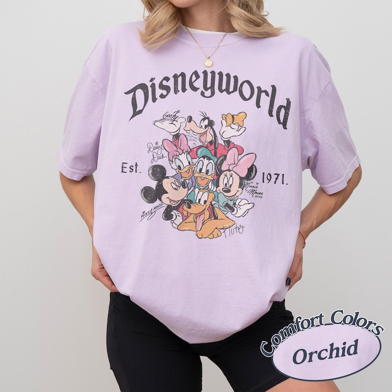 Może przedstawiać: Jasnofioletowa koszulka z napisem "Disneyworld" w ozdobnej czcionce, z grafiką postaci Disneya. Koszulka zawiera napis "Est. 1971." i logo Comfort Colors Orchid.
