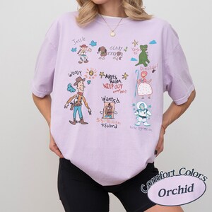 Puede incluir: Una camiseta Comfort Colors de color lila claro con ilustraciones dibujadas a mano de personajes de Toy Story. El dise&ntilde;o incluye a Woody, Buzz Lightyear, Jessie y Slinky. El texto incluye "Andy's Room Keep Out" y "Wanted $50 Billion Reward."