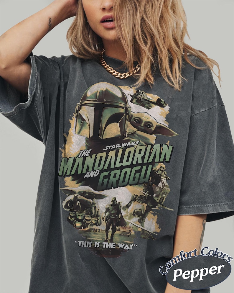 Może przedstawiać: Ciemnoszara koszulka z grafiką z Gwiezdnych Wojen: The Mandalorian. Projekt zawiera hełm Mandalorianina, Grogu i tekst "THE MANDALORIAN AND GROGU". Na koszulce wydrukowano r&oacute;wnież frazę "THIS IS THE WAY".