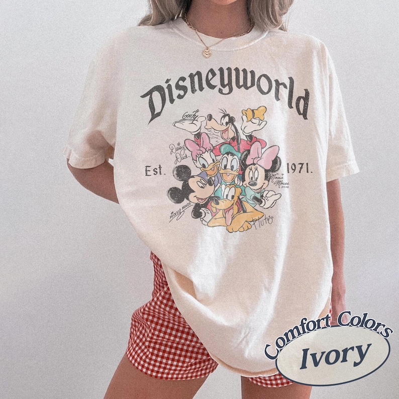 Może przedstawiać: Kremowa koszulka z grafiką Disneyworld z Myszką Miki, Myszka Minnie, Kaczorem Donaldem, Daisy, Goofym i Plutonem. Koszulka zawiera napis "Disneyworld Est. 1971". Marka Comfort Colors.
