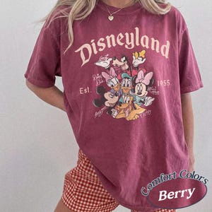 May include: Berry-colored Disneyland t-shirt featuring a graphic of Mickey Mouse, Minnie Mouse, Donald Duck, Daisy Duck, Goofy, and Pluto. The shirt has the text "Disneyland" and "Est. 1955." The shirt is a Comfort Colors brand.